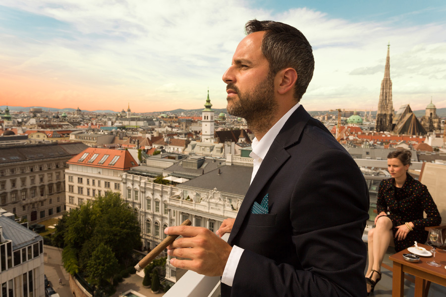Ein Mann steht beim Rauchen und schaut über die Stadt Wien hinaus und eine Frau sitzt und raucht in einem Stuhl